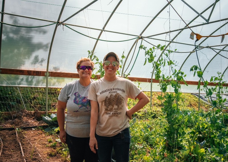 Two women standing in a high tunnel with green plants.