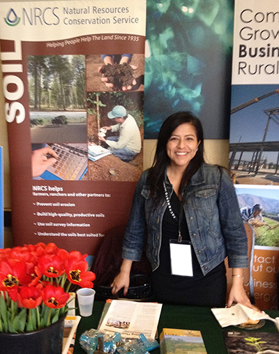 Christine Chavez, a woman with black hair, wearing a jean jacket and a black dress, standings behind a table holding NRCS publications and a bucket of red flowers
