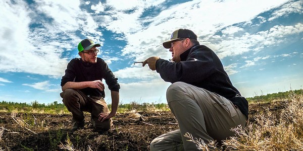 Two men wearing baseball caps crouch in a field as they talk