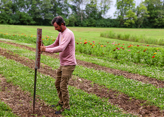Tyler Gough, a white man in a pink shirt and brown pants, handles farm equipment in a field.