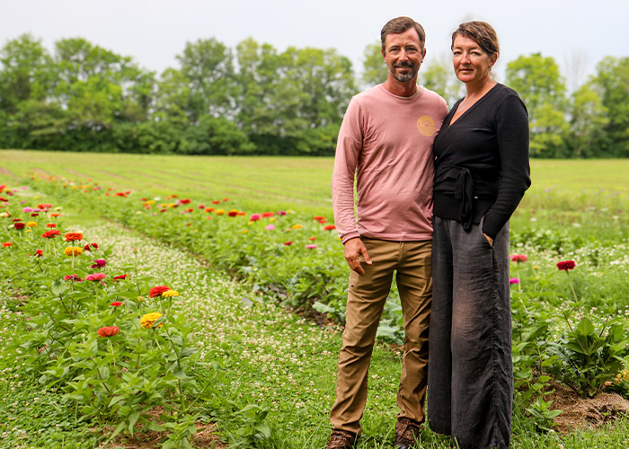 A man and woman stand next to each other on a field with rows of flowers planted behind them.
