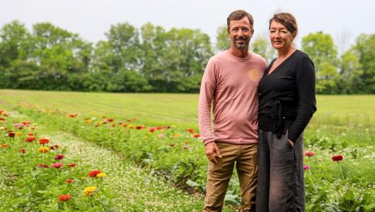 A man and woman stand next to each other on a field with rows of flowers planted behind them.